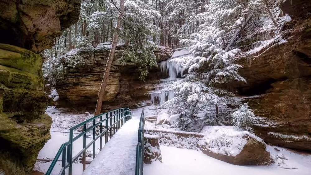 Hocking Hills Snow-covered footbridge leading toward a partially frozen waterfall surrounded by rocky cliffs and winter forest